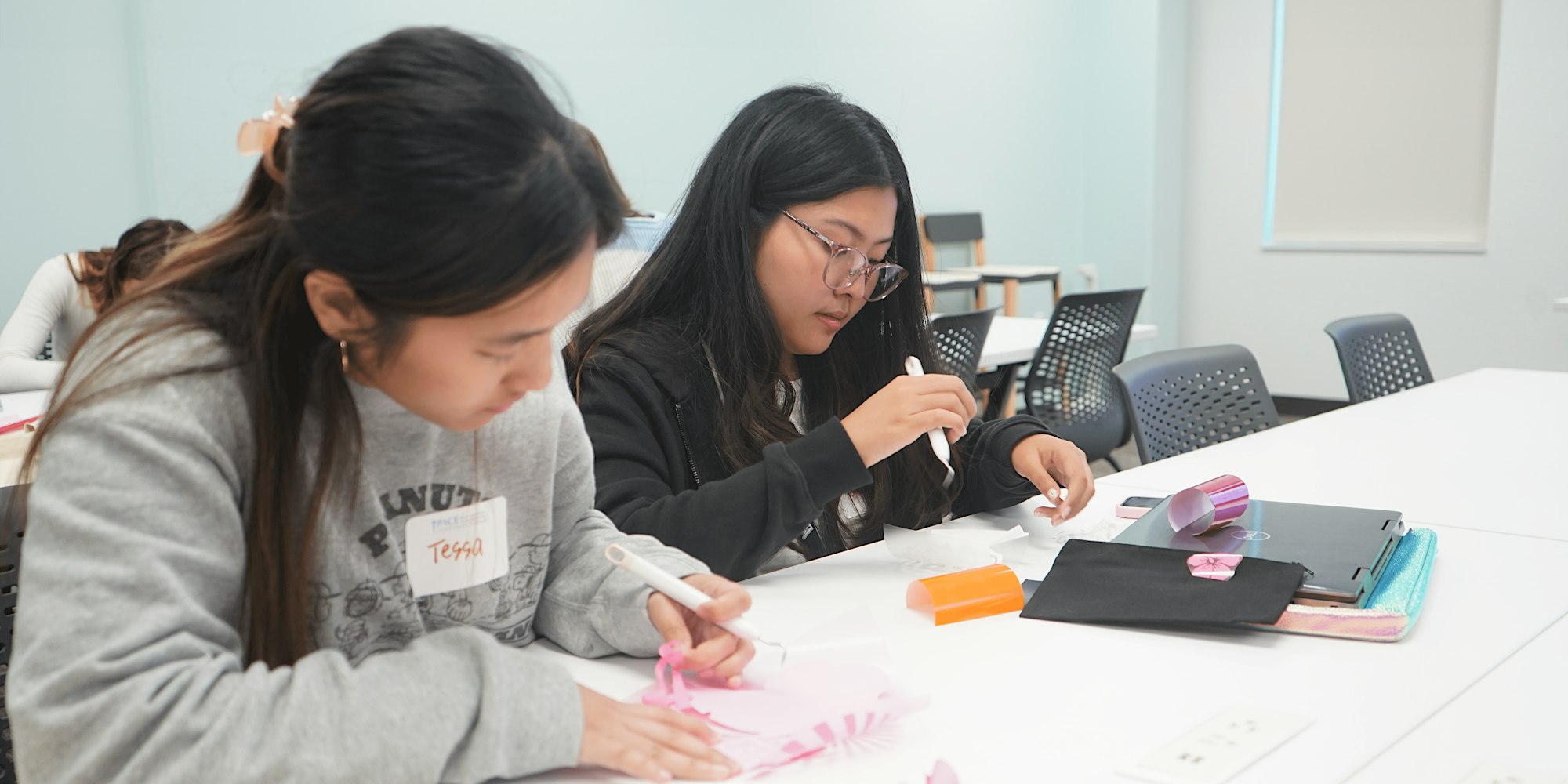 Two girls sitting at a white table peeling adhesive to make a design at a PACE Make and Take event